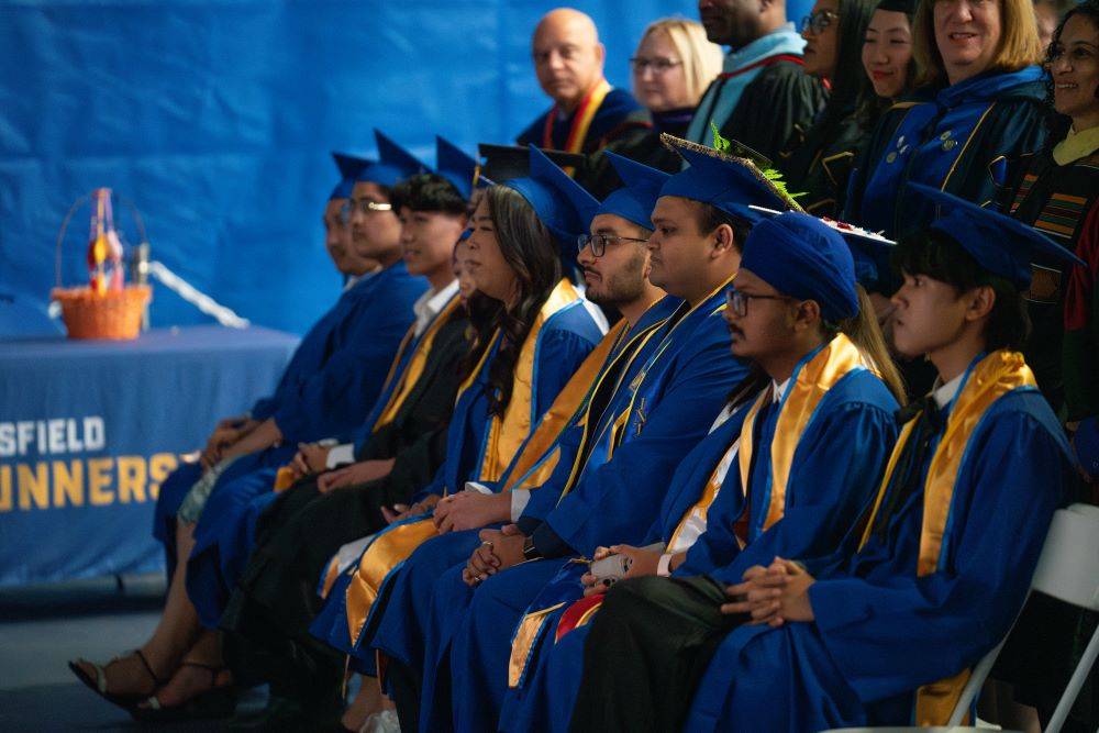 Asian American Pacific Islander Desi American (APIDA) Graduation Spring 2025 - CSUB Students dressed in blue gowns and caps with white and gold stole sit in a line while faculty and staff stand behind them.