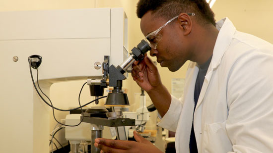 Male student in safety glasses and lab coat looking through microscope