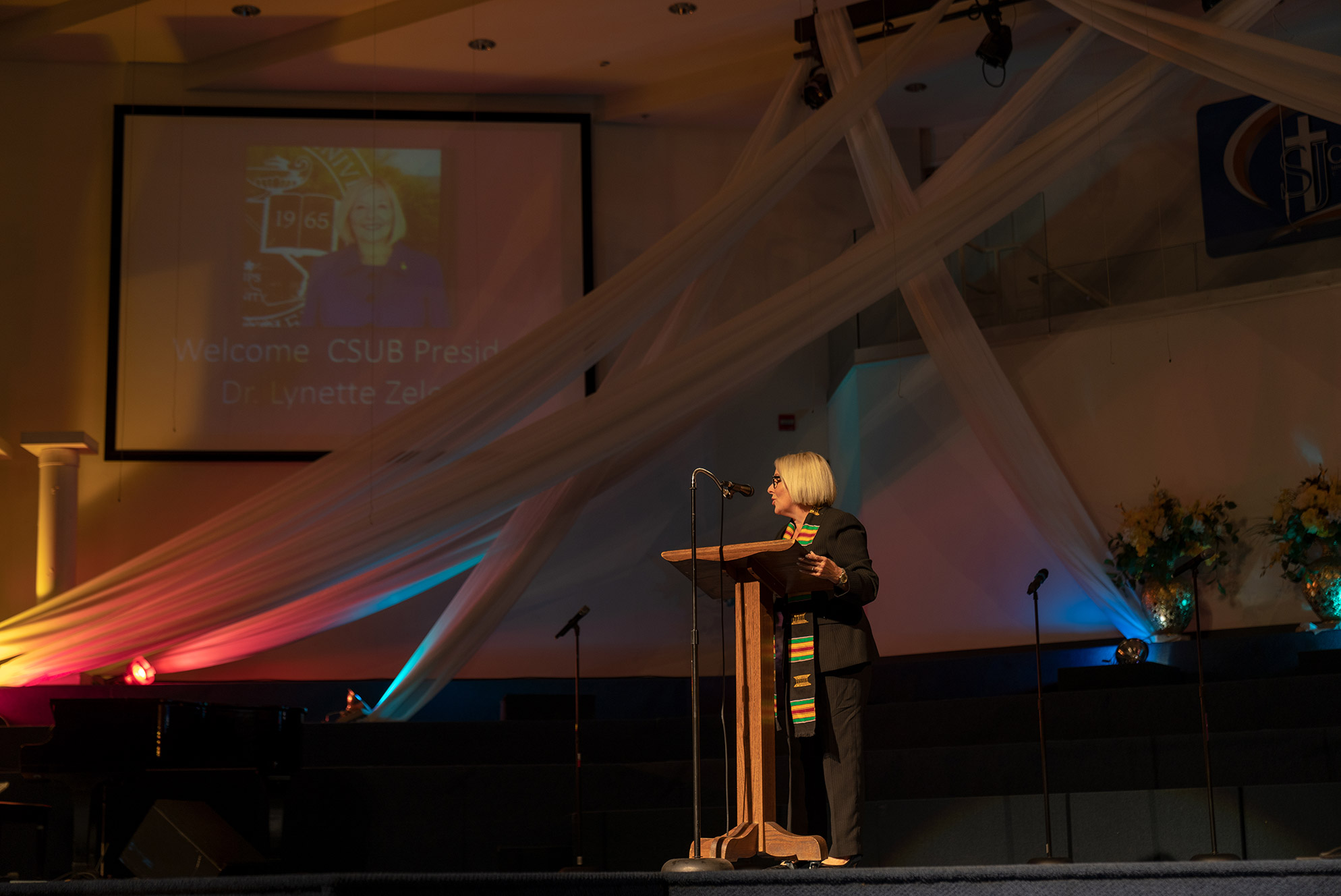Woman on stage and speaking into microphone with large video screen in background