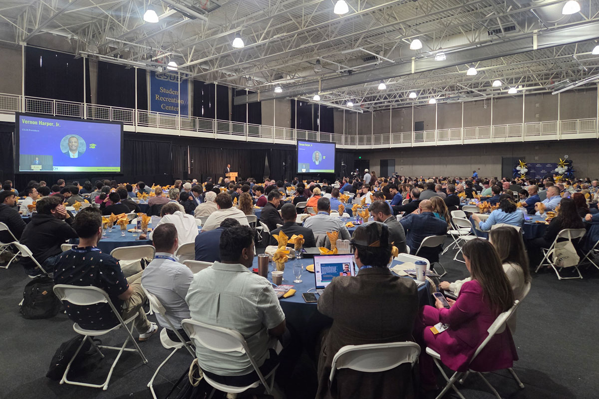 Attendees seated at round tables listen to a speaker presenting at a podium during a large CSUB conference.