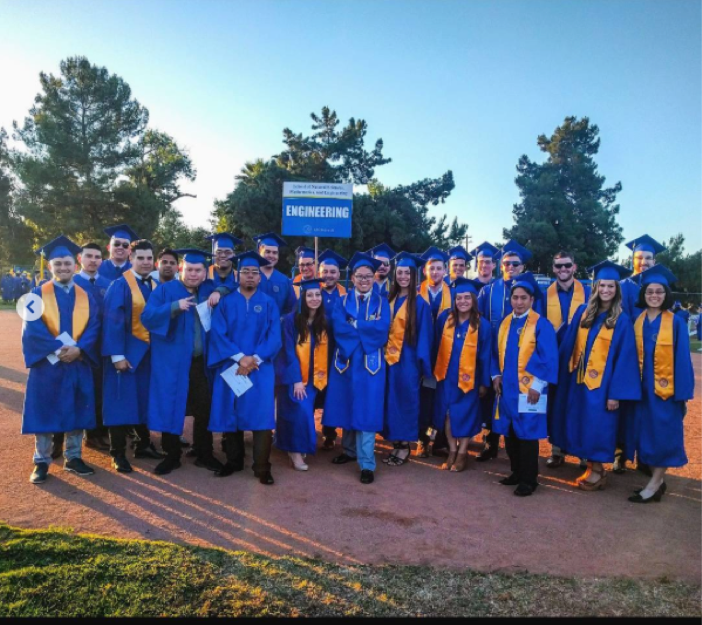 A group of engineering graduates in blue gowns pose together outdoors holding an 'Engineering' sign