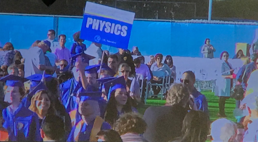 Physics graduates in caps and gowns walk in a commencement procession holding a 'Physics' sign