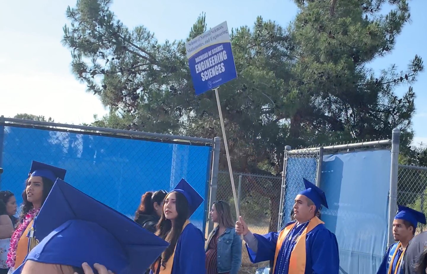 Engineering graduates in blue gowns walk in a commencement procession holding a department sign