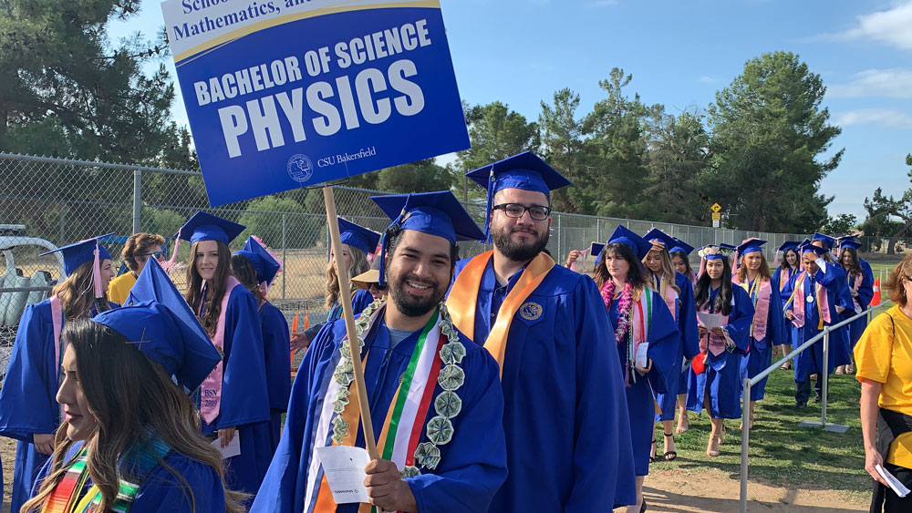 Physics graduates in blue caps and gowns walk together during a commencement ceremony holding a 'Physics' sign